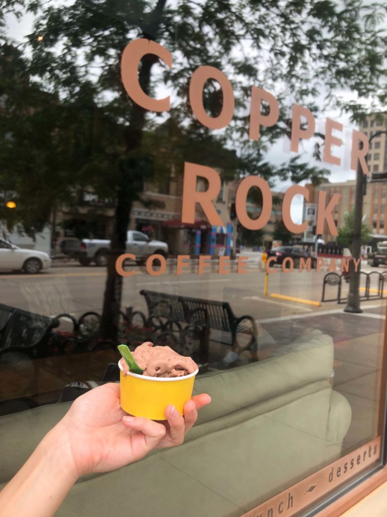 A hand hold a cup of chocolate ice cream in front of the Copper Rock store front.